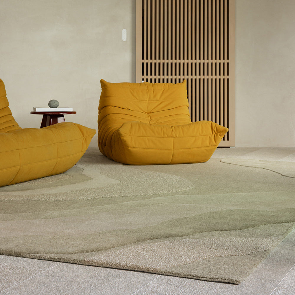 A mustard yellow modular lounge chair set in a minimalist, warm-toned room with a textured beige rug, a wooden slatted screen, and a small side table.