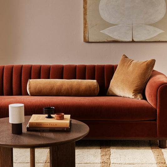 A warm rust-red velvet sofa in a styled living room setting, decorated with a tan cylindrical lumbar pillow and a square tan throw pillow, set against a beige wall with abstract wall art.