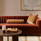 A warm rust-red velvet sofa in a styled living room setting, decorated with a tan cylindrical lumbar pillow and a square tan throw pillow, set against a beige wall with abstract wall art.