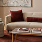 A styled living room scene featuring a beige textured boucle sofa with deep burgundy velvet throw pillows, including a cylindrical lumbar pillow, set against a warm beige wall with a large abstract canvas print.
