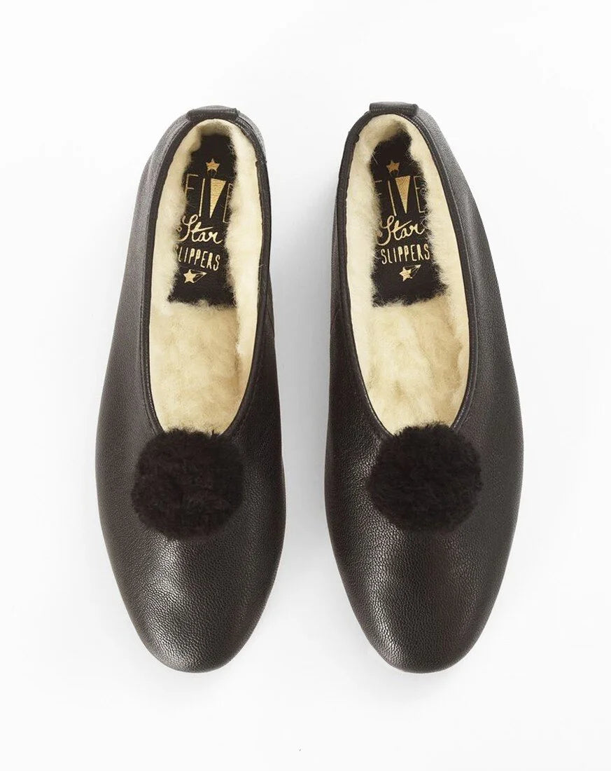 A pair of black leather slip-on flats with fluffy black pom-poms on the toe and cream-colored shearling lining, shown from a top-down view on a white background.