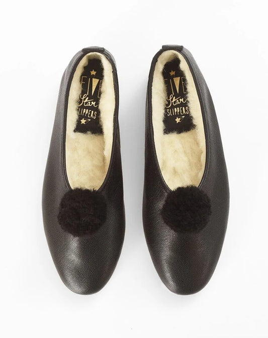 A pair of black leather slip-on flats with fluffy black pom-poms on the toe and cream-colored shearling lining, shown from a top-down view on a white background.