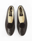 A pair of black leather slip-on flats with fluffy black pom-poms on the toe and cream-colored shearling lining, shown from a top-down view on a white background.