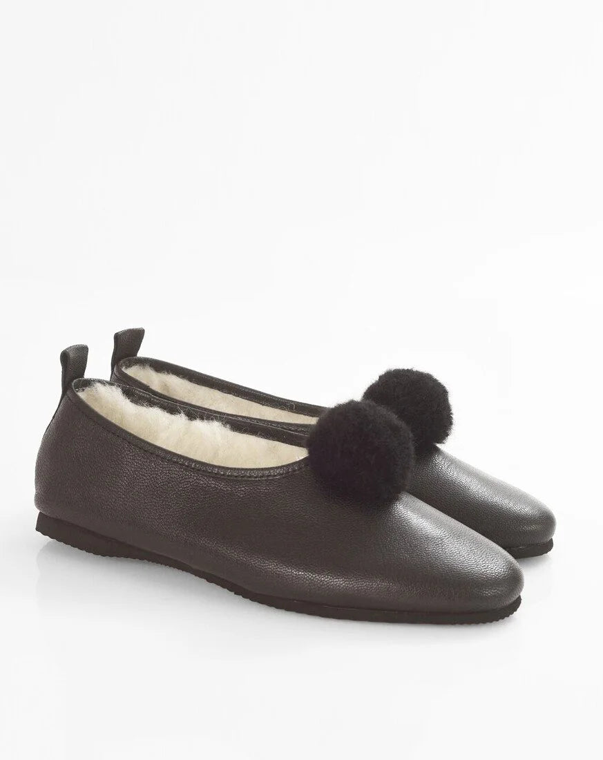 A pair of dark brown leather ballet flats with fluffy black pom-poms on the toes and a cream-colored shearling lining, shown against a plain white background.