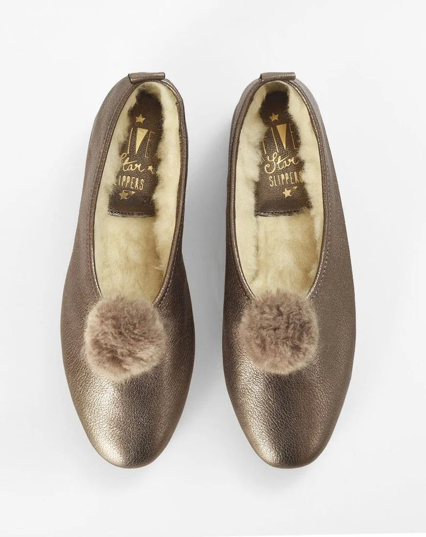 A pair of metallic bronze slip-on flats with fluffy faux-fur pom-poms on the toe and a cream-colored shearling lining, shown from a top-down view on a white background.