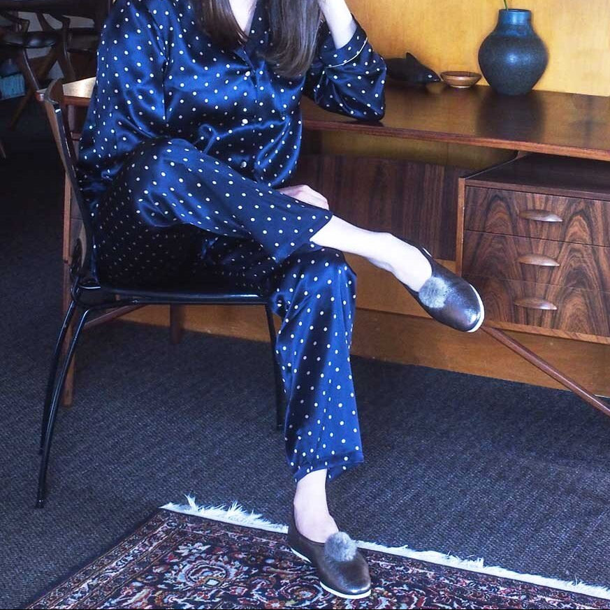 A woman wearing a navy blue pajama set with white polka dots sits on a chair in a home interior setting.