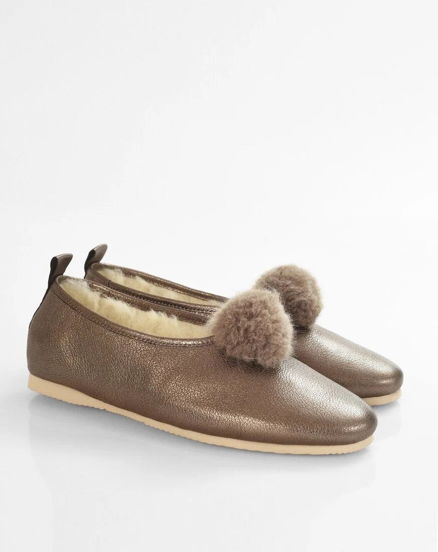 A pair of women's slip-on flats in metallic bronze leather with fluffy brown faux-fur pom-poms on the toe and a cream shearling lining, shown on a white background.