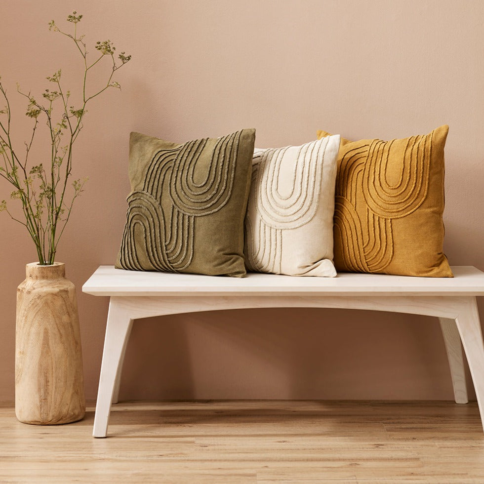 Three textured Gatsby cushions in toffee, olive and ecru sitting on a pale wooden bench with a peach wall and wooden vase with dried flowers on the wooden floor beside the bench