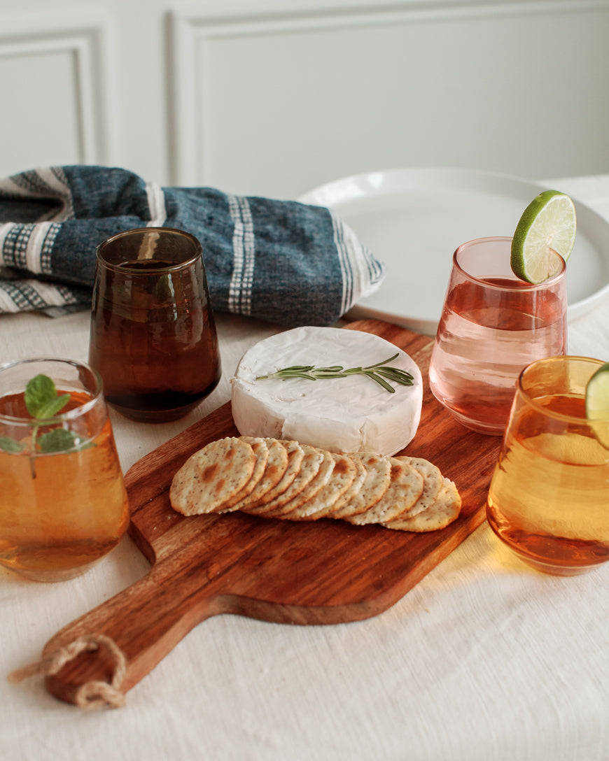 Wooden cutting board with cheese, crackers, and glasses of tea on a white tablecloth.