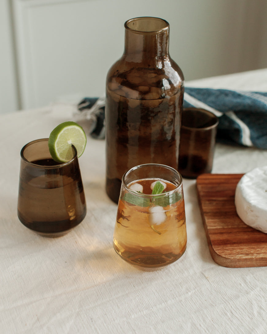 Two glasses of iced tea with a lime wedge and mint leaf, accompanied by a brown carafe on a light surface.