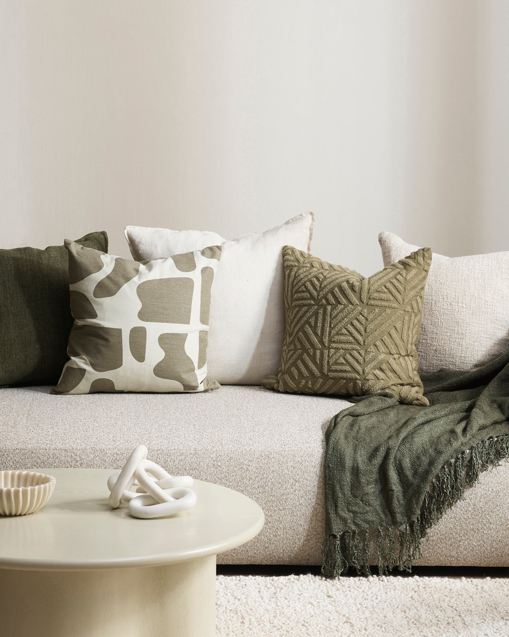 Harper cushion with cream back ground and abstract olive sploges from the Baya Collection in lifestyle shot with other cushions, cream sofa and rug,