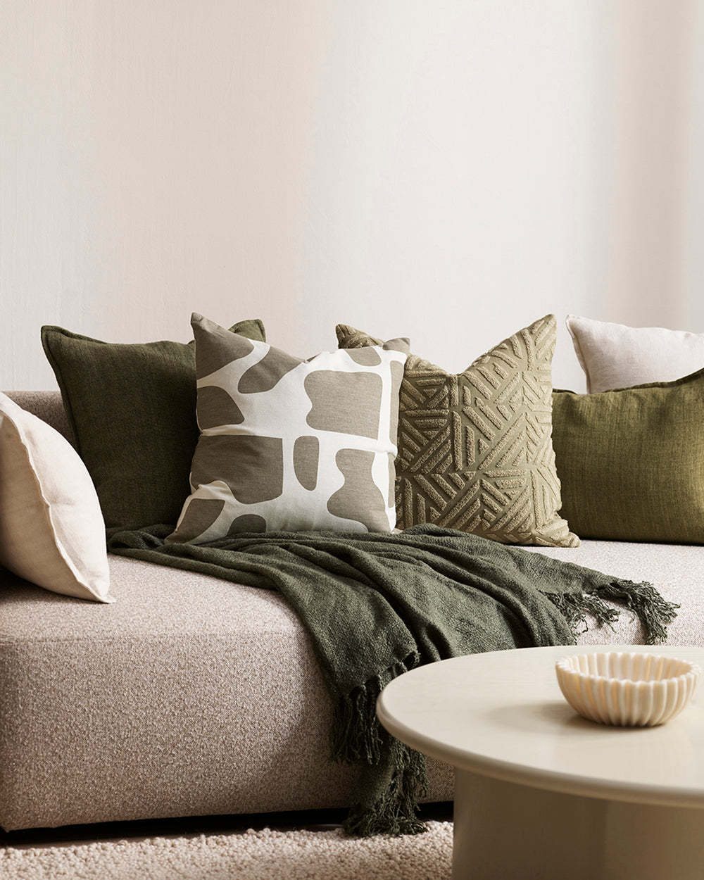 Harper cushion with cream back ground and abstract olive sploges from the Baya Collection in lifestyle shot with other cushions, cream sofa and rug,