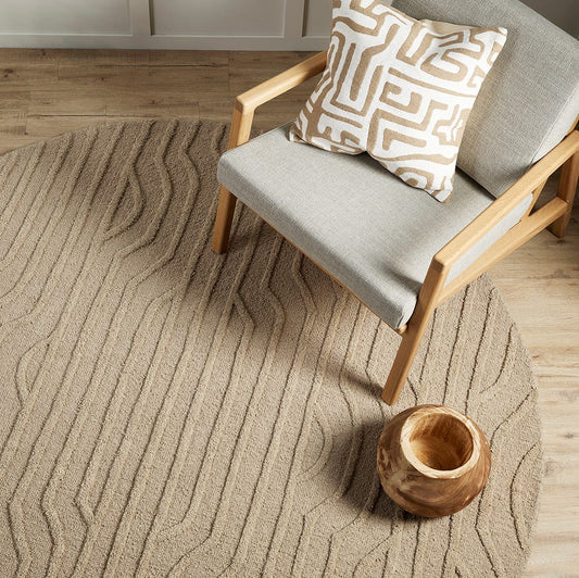 round wool rug in pebble with textured art deco type lines with a grey arm chair and cream and beige abstract patterned cushion on top