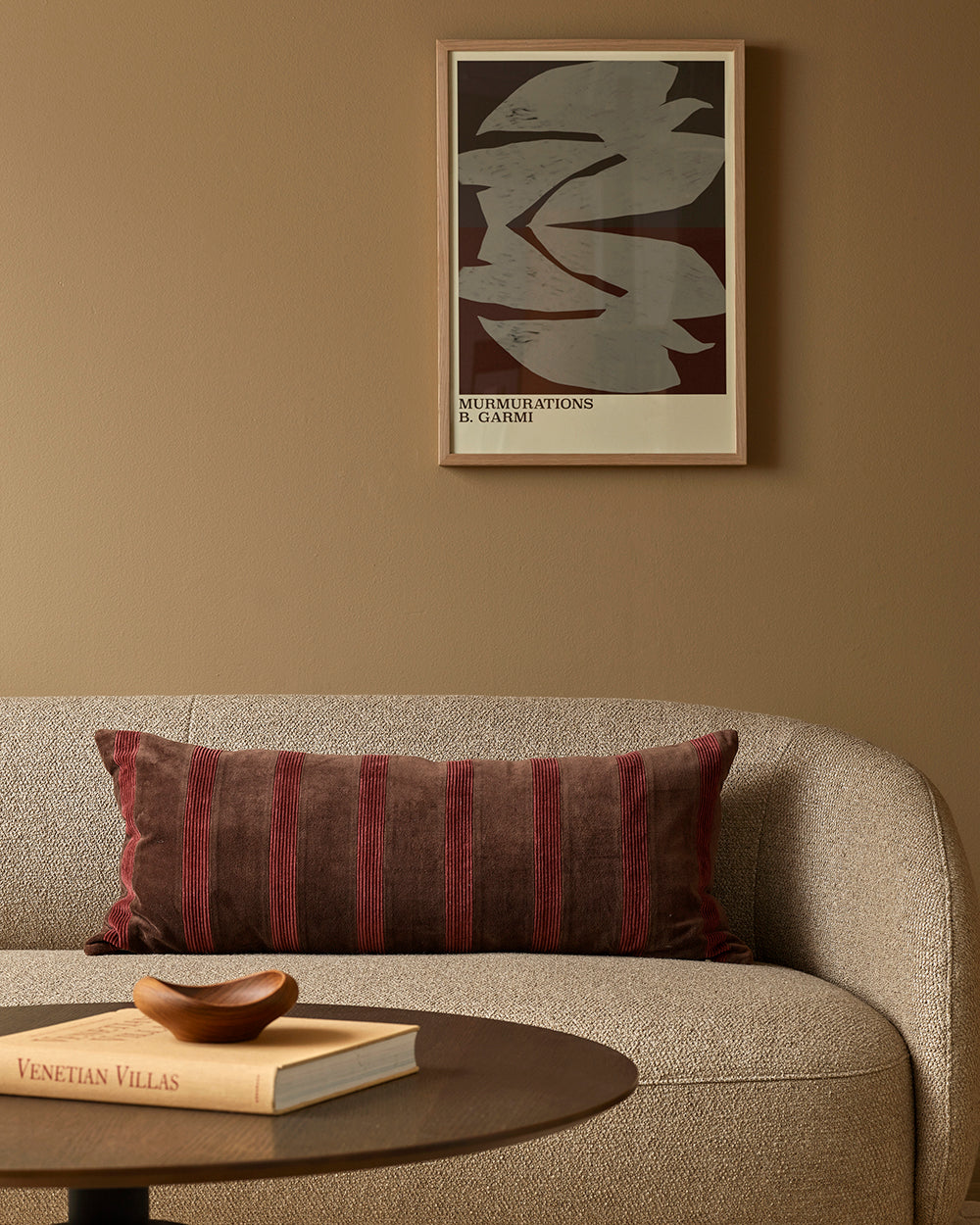 Sofa with striped pillow, wooden bowl, and book on a small table against a beige wall with a framed abstract art piece.