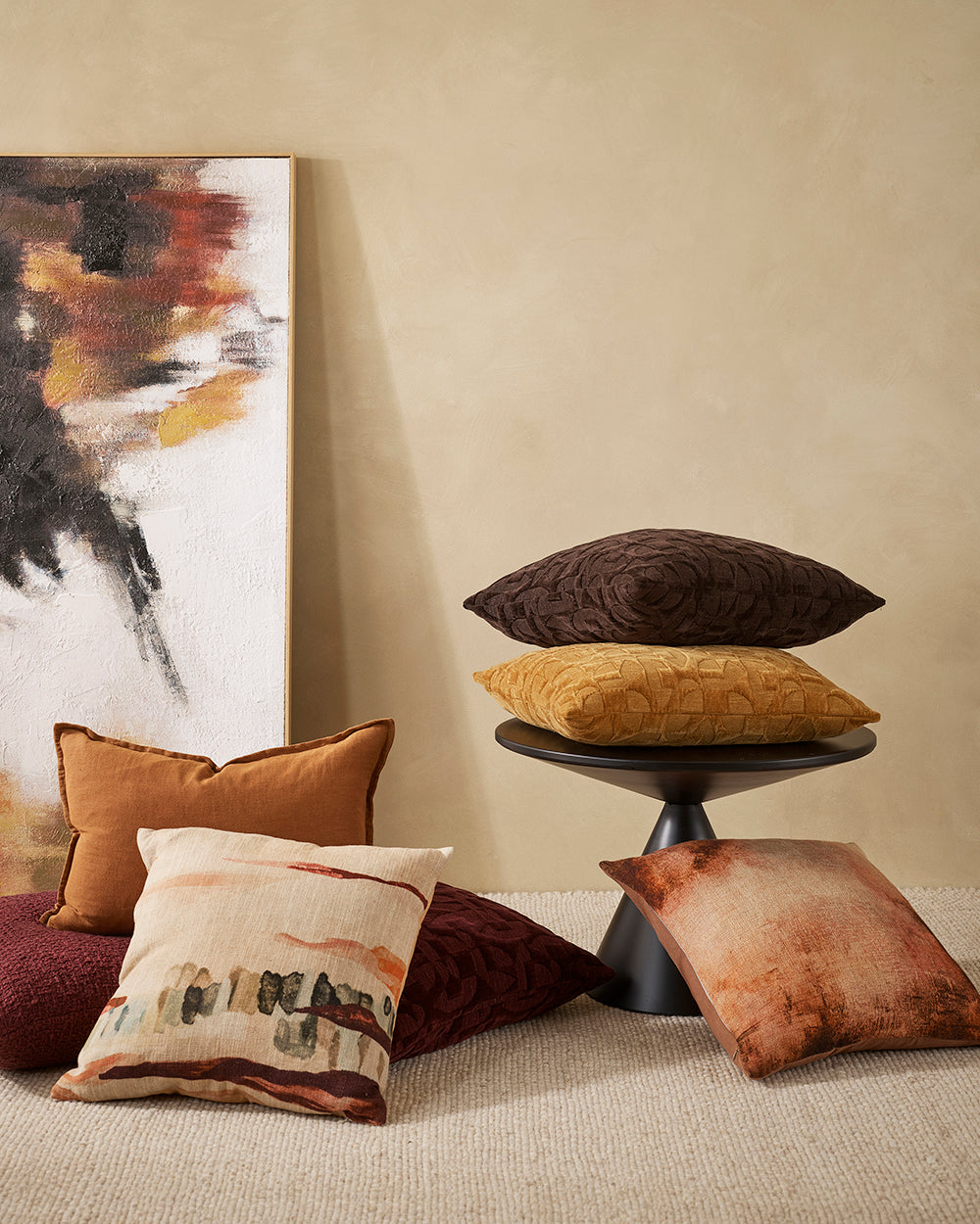 mix of cushions in pomegranate, toffee and chocolate, one on the floor, 2 on a black modern round side table with a painting leaving against a cream wall in gold, charcoal, chocolate, orange
