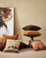 mix of cushions in pomegranate, toffee and chocolate, one on the floor, 2 on a black modern round side table with a painting leaving against a cream wall in gold, charcoal, chocolate, orange