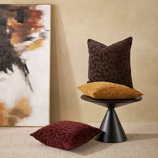 jacquard cushions in pomegranate, toffee and chocolate, one on the floor, 2 on a black modern round side table with a painting leaving against a cream wall in gold, charcoal, chocolate, orange