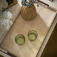 Two green glass tumblers on a textured surface with a woven container and white flowers in the background.