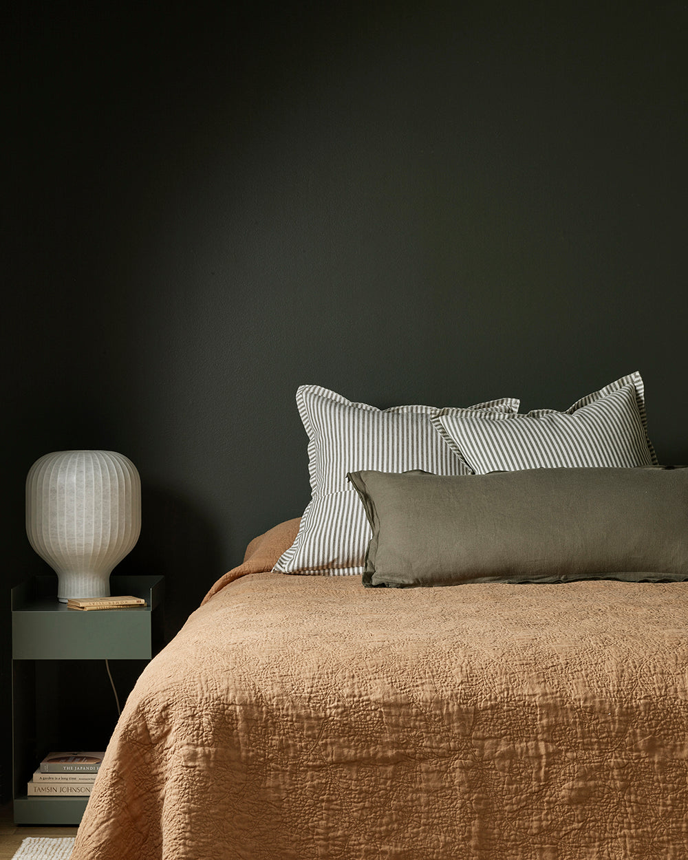 Bed with brown bedding and striped pillows against a dark wall, with a nightstand and lamp.