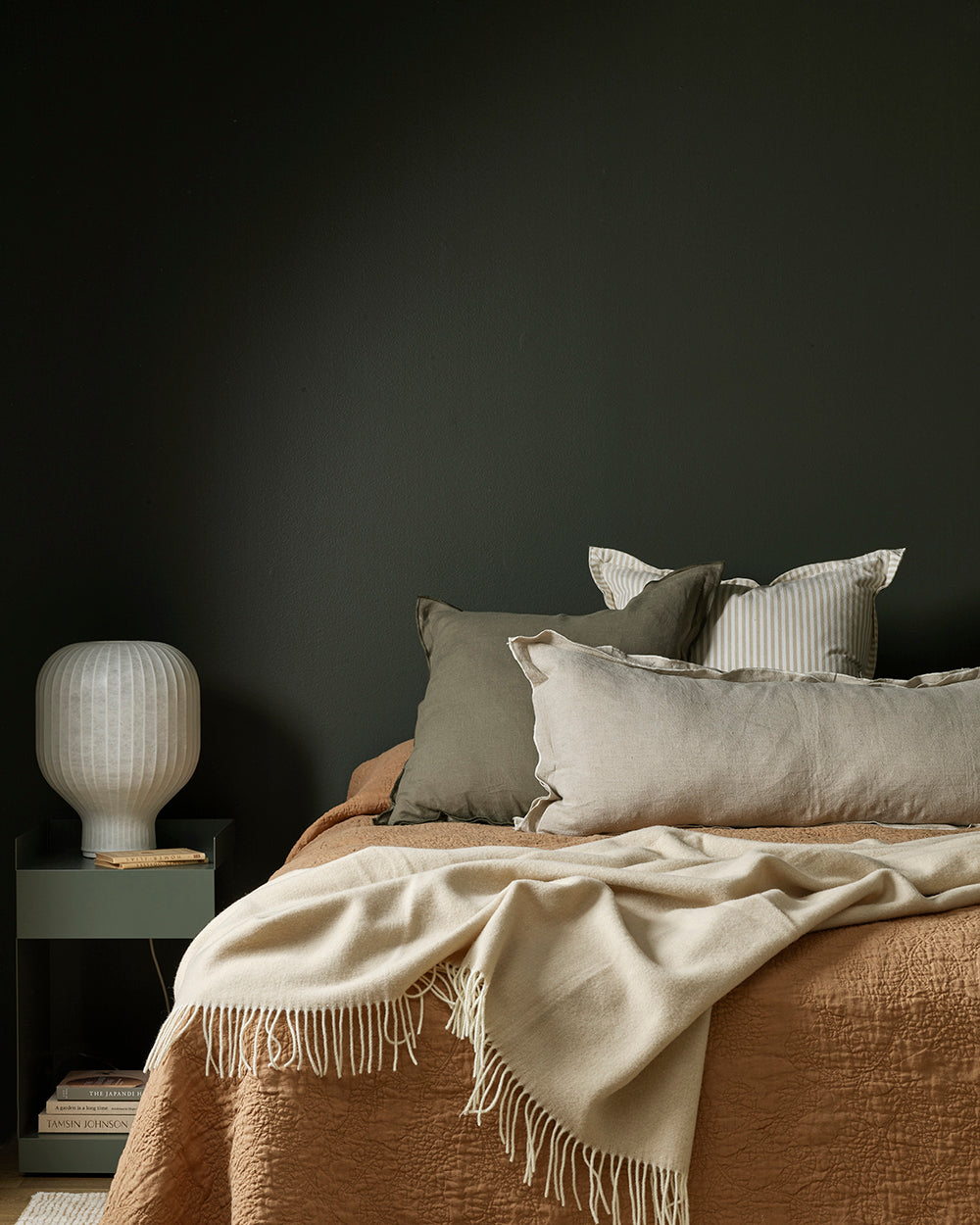 Bed with beige bedding and pillows against a dark wall with a side table and lamp.