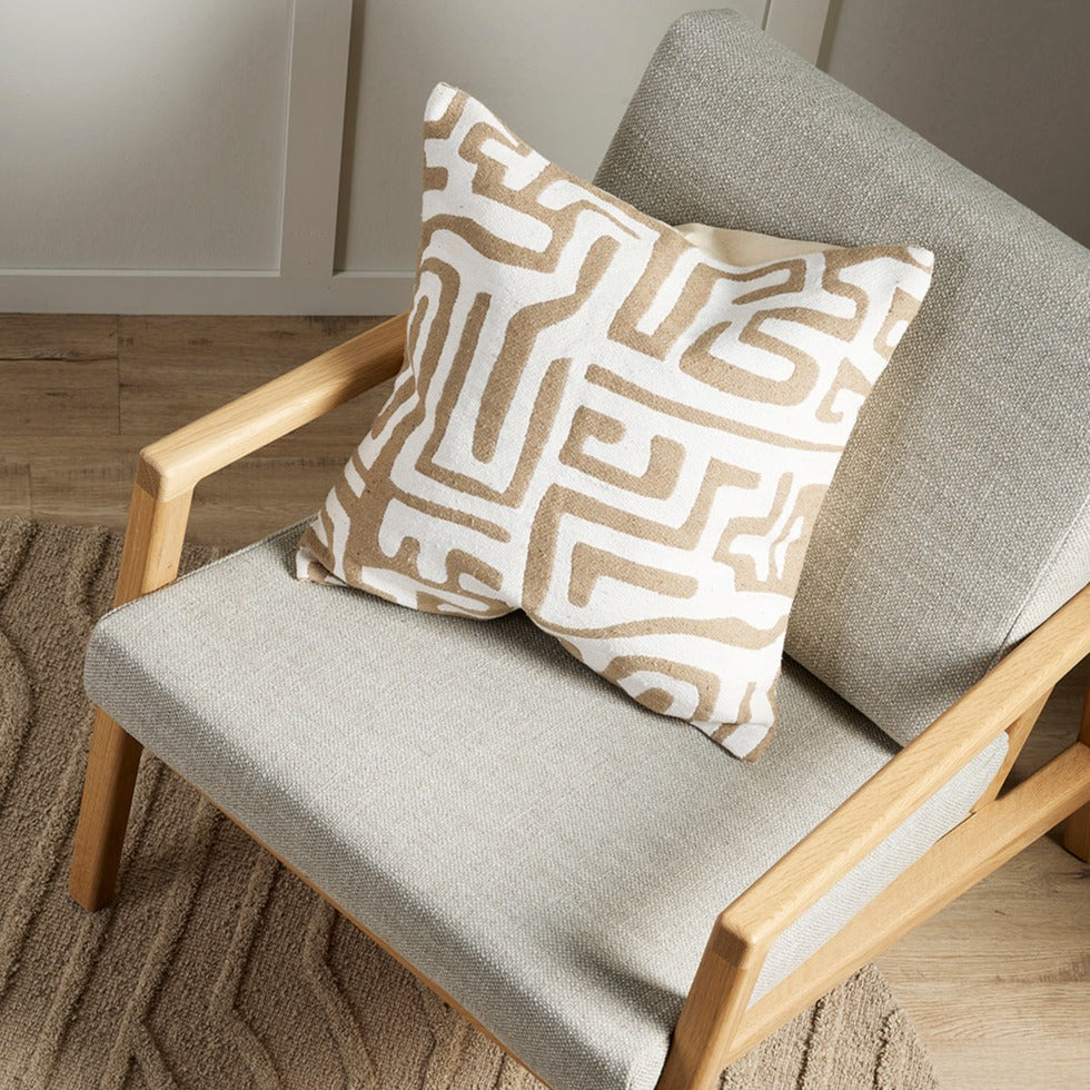 natural and white geometric pattern on cushion sitting on a grey mid century style wooden chair on a beige textured rug on a wooden floor with wainscotting just seen on the walls