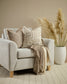 Beige armchair with patterned cushions and a blanket, accompanied by a vase with dried plants on a wooden floor.
