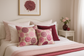 Magazine-style feminine bedroom featuring pink chrysanthemum cushions on white bed with pale pink linens and burgundy accents