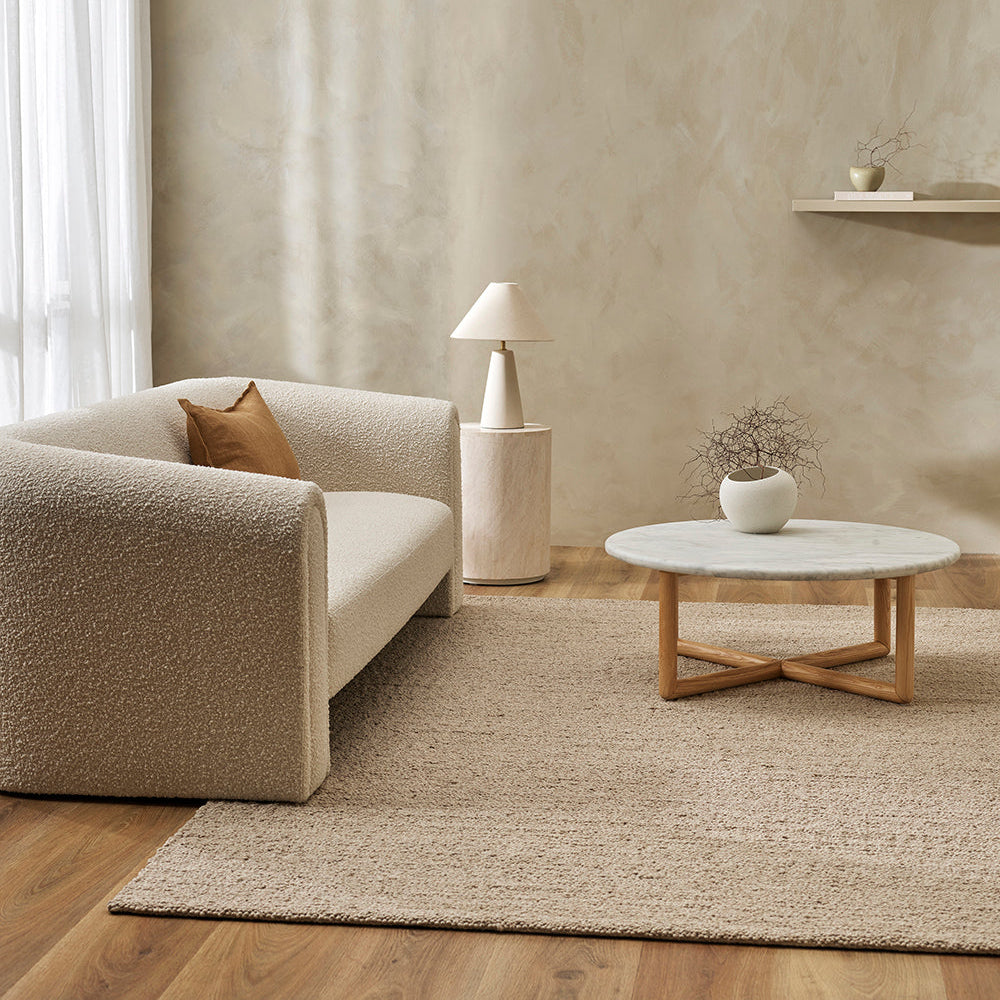A beige wool rug with a textured design, placed on a wooden floor in a living room setting, complementing a neutral color palette decor.
