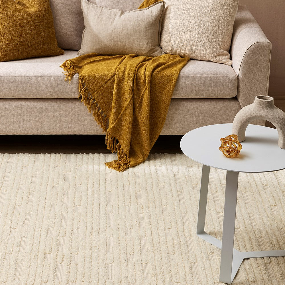 Myriam rug in ecru with linear tufted design. a cream cushion is set on the rug with a mixture of ecru and toffee colour cushions with a toffee throw draped on the sofa. A metal white coffee table is in the forefront with an amber class cage ball object and a stone arched object.