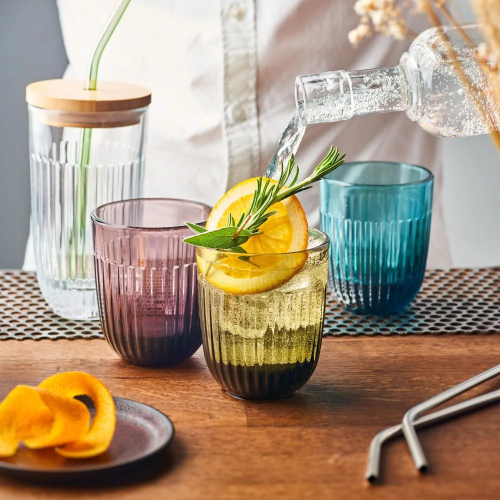 Glass of lemonade with a slice of lemon and rosemary, surrounded by other glasses and a bottle on a wooden table.
