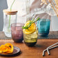 Glass of lemonade with a slice of lemon and rosemary, surrounded by other glasses and a bottle on a wooden table.