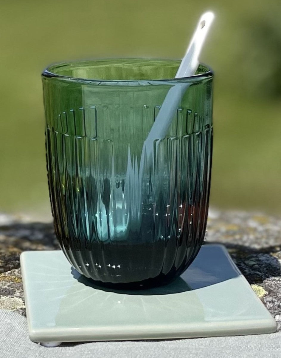 A dark blue ribbed glass tumbler with a white plastic spoon inside, resting on a square light green coaster outdoors.