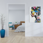 A colorful abstract portrait canvas print of a woman's face in profile, featuring vibrant blue, yellow, pink, and black brushstrokes on a white background, displayed on a white wall in a modern bedroom setting.