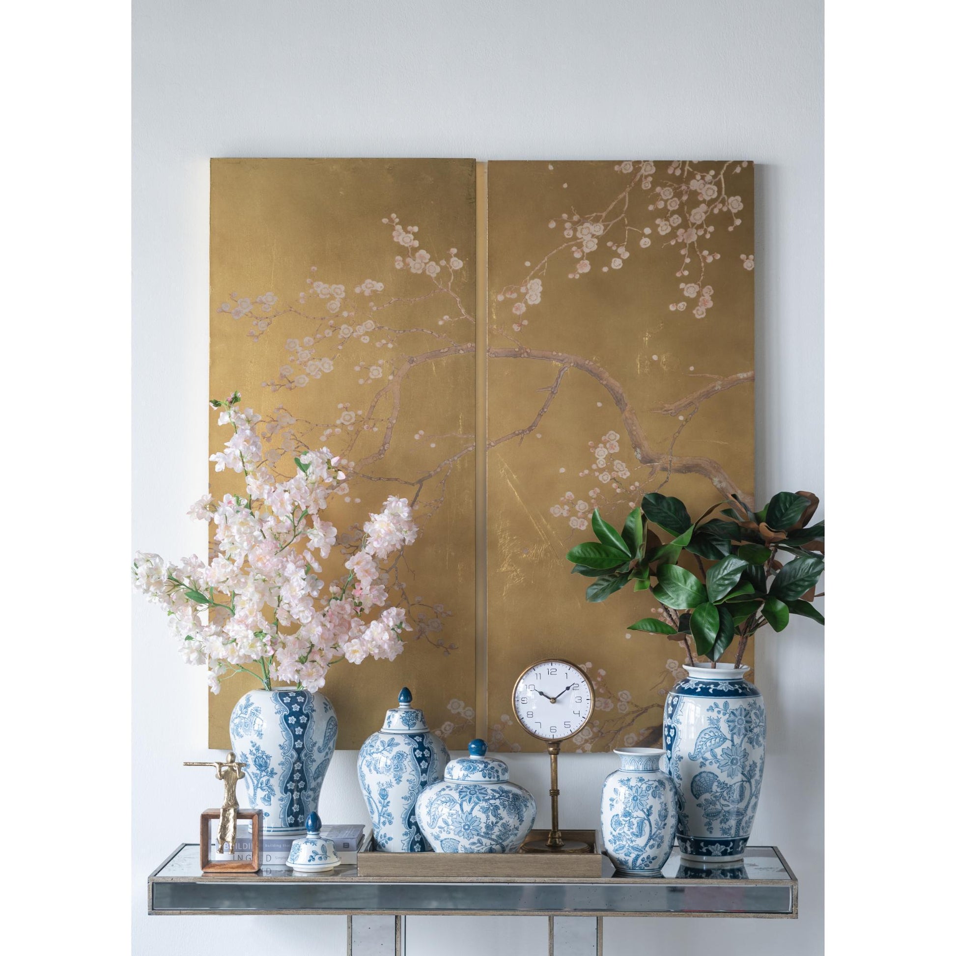 Decorative setup with vases, a clock, and a plant against a floral wall painting.