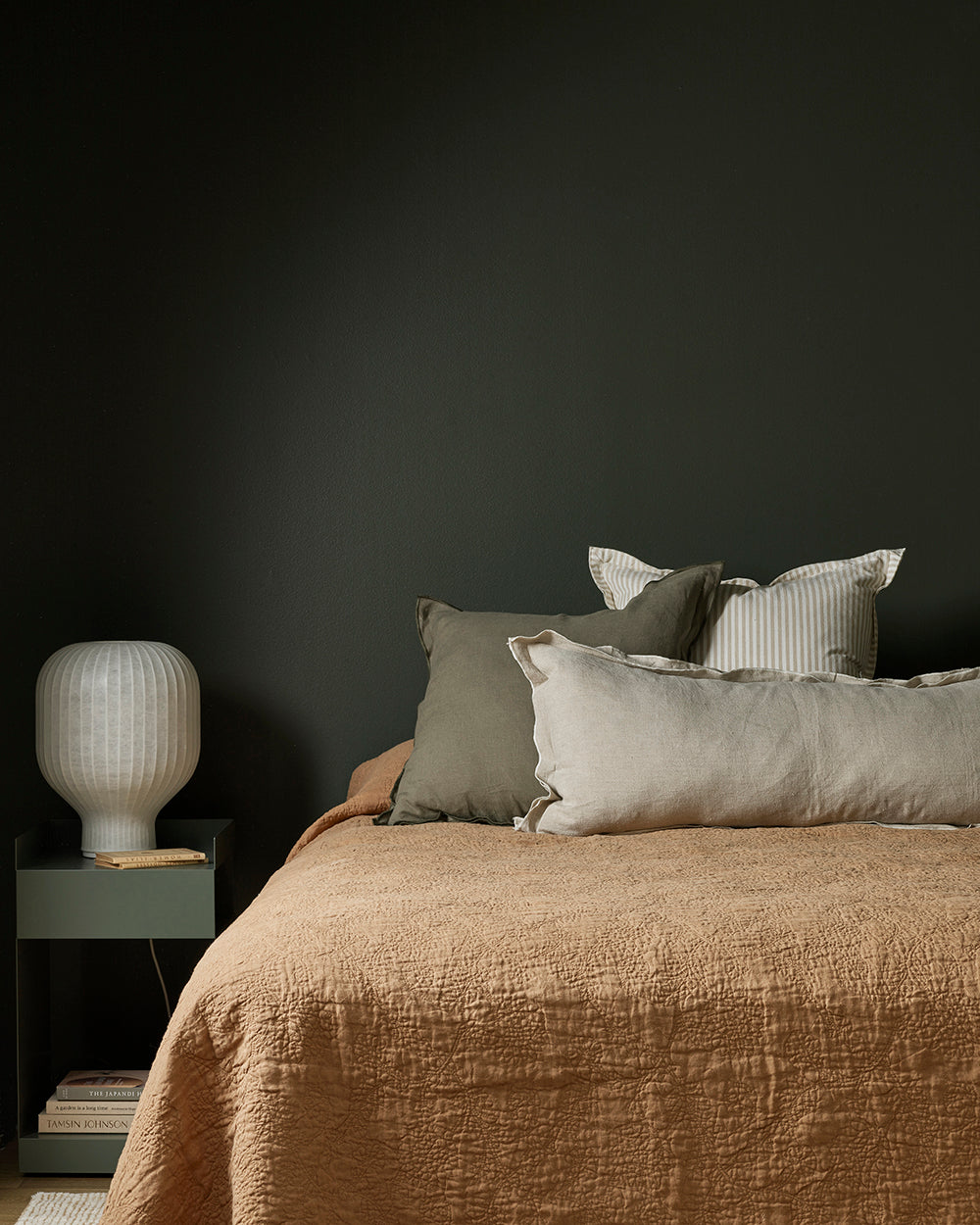 Bed with beige bedding and pillows against a dark wall with a nightstand and lamp.