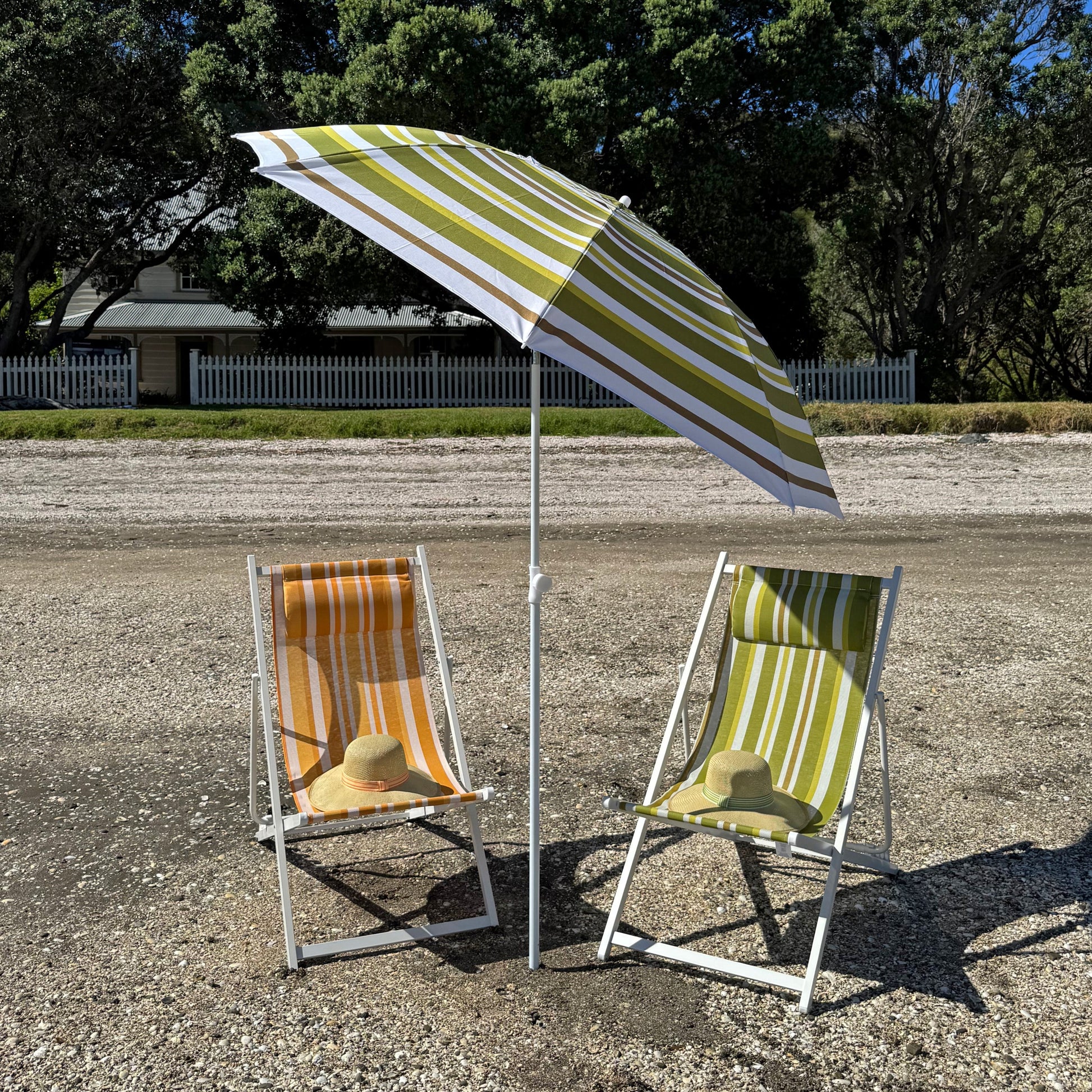 Two beach chairs under a striped umbrella on a gravel surface with trees in the background.