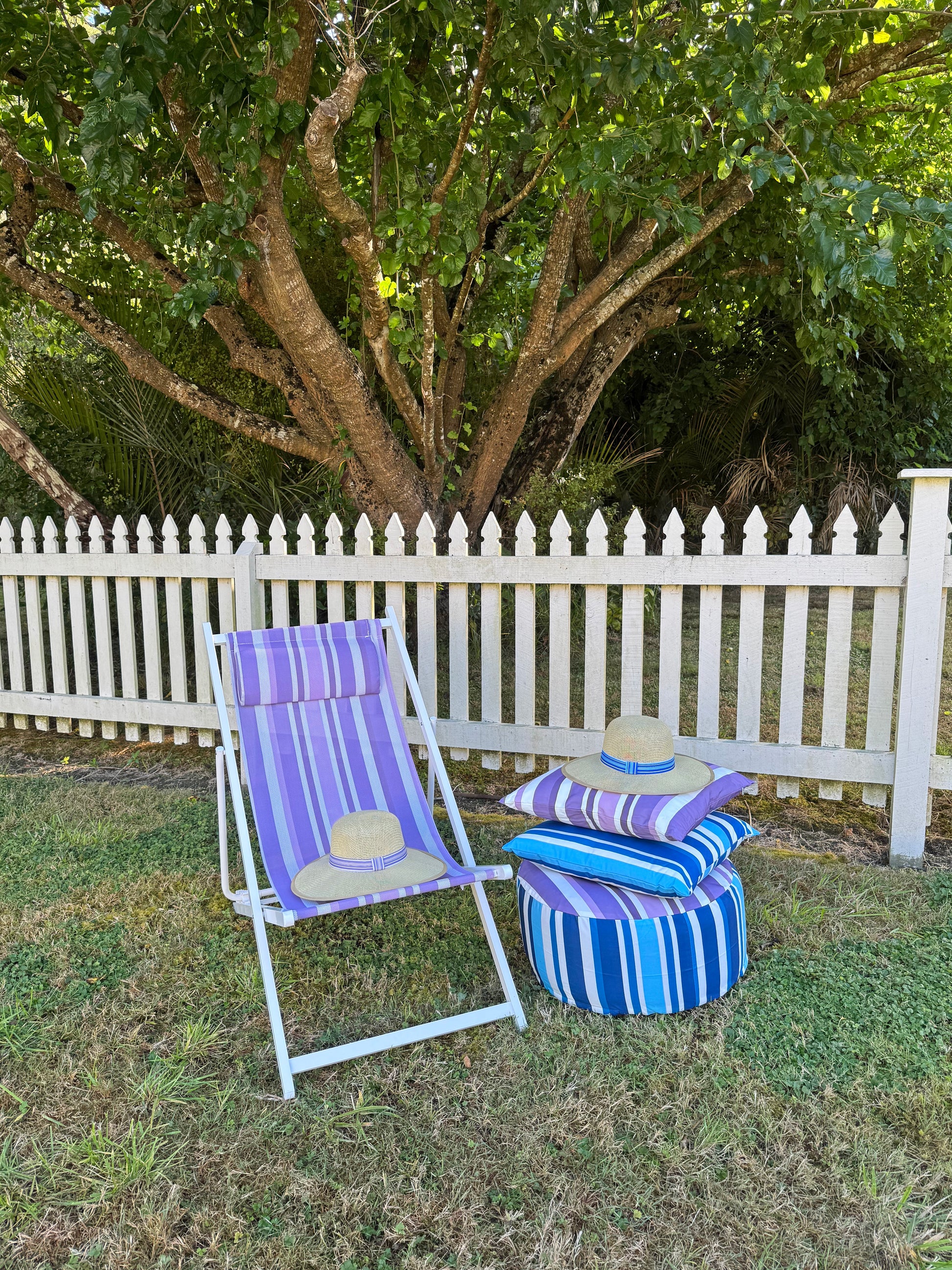 Outdoor setting with a striped deck chair, ottoman, and pillows against a white picket fence.