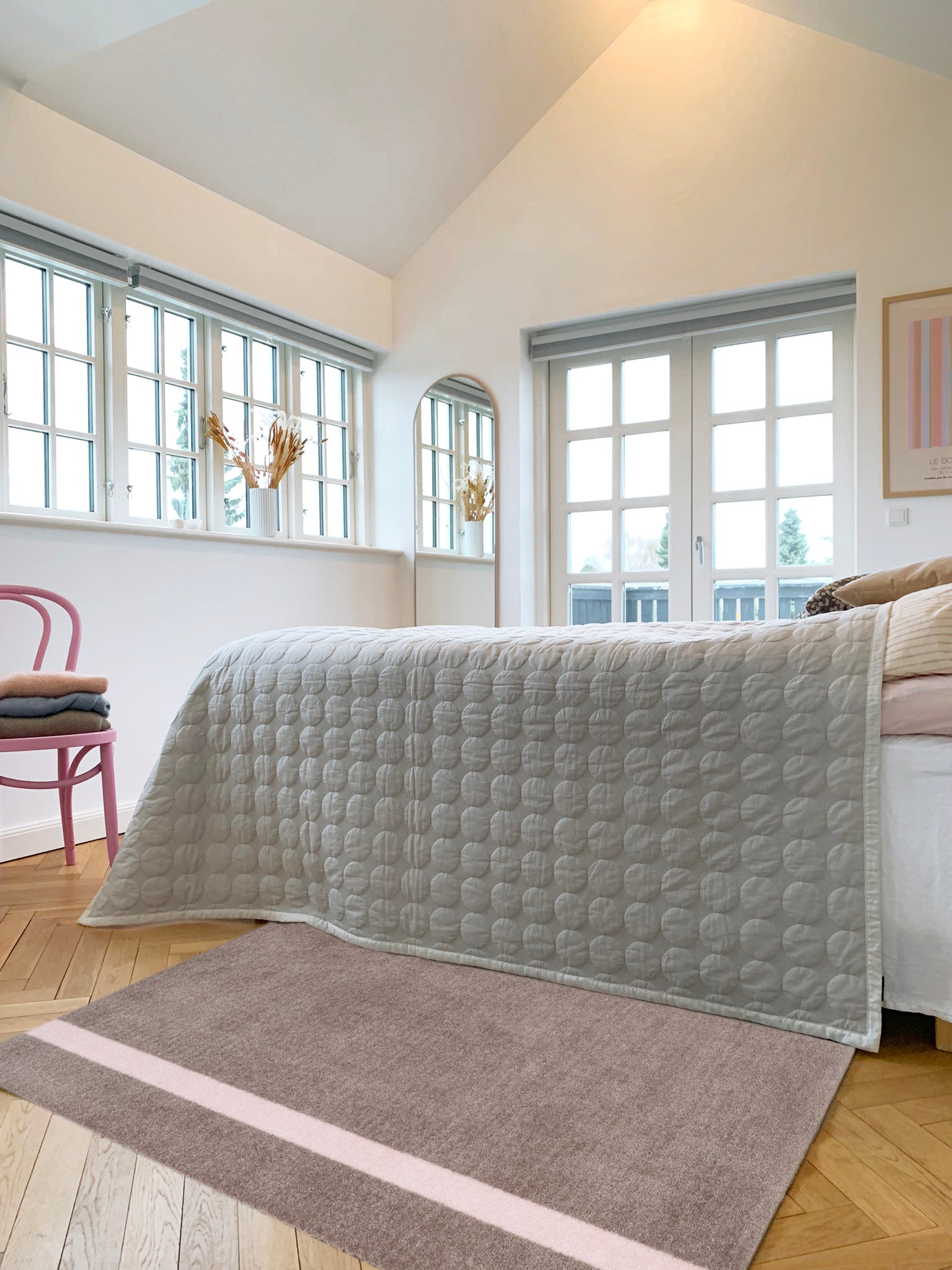 Gray quilt draped over a bed in a bright room with large windows.