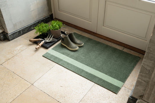 Green doormat with striped pattern on a tiled floor, next to a pair of boots and gardening tools.