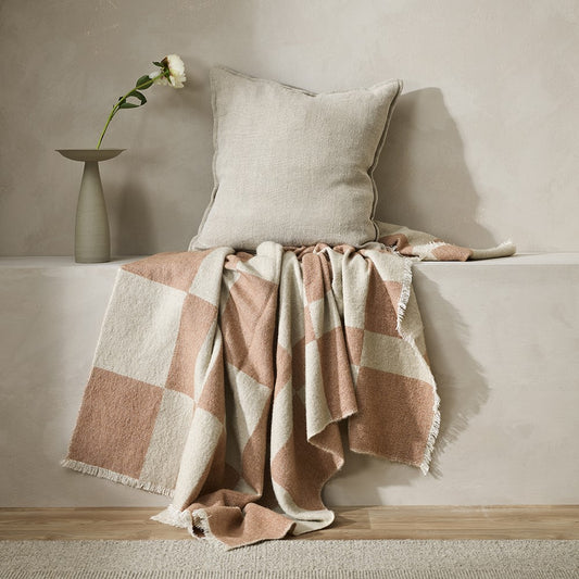 Winton throw in a coral and neutral check draped against a stone wall alcove with a cream linen cushion on top. Also a gren modern vase with a white carnation in