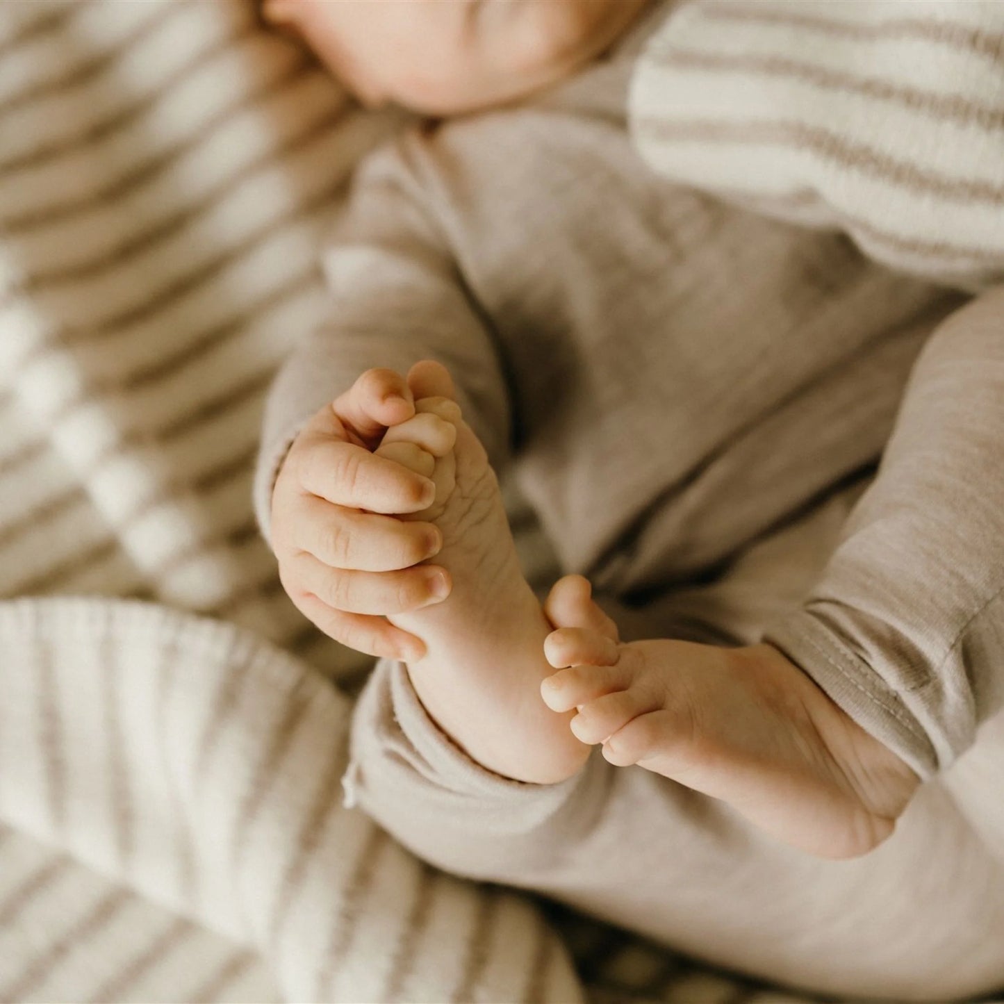 Close-up of a baby's feet with toes intertwined, wearing a beige outfit on a soft textured background.