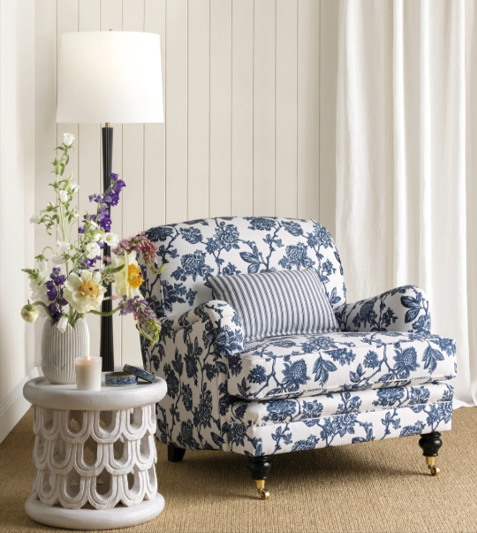 Fleur fabric in botanical motifs on sofa with a white morrocan style side table with a vase of yellow and purple flowers and a tall white shaded lamp with a black stem