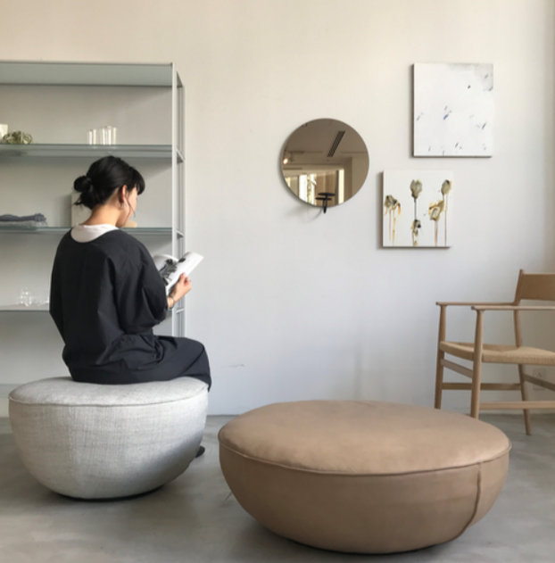 Natadora fable leather ottoman in lifestyle shot with woman sitting on the fabric version in a lounge with a wooden chair and metal shelving
