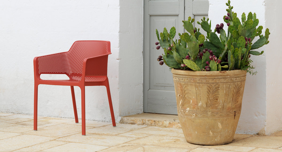 nardi net chair in coral red outside a meditteranean grey door, white walls and a rustic plant pot with cactus in fruit