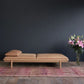 Tolv pensive day bed in light tan leather in front of a dark blue wall and on a pink rug with a vase of pink lillies