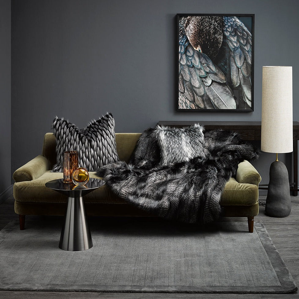 Alaskan Wolf imitation fur throw from heirloom in a lifestyle shote, grey scene with bird picture on the wall and Alaskan Wolf cushion on a green velvet sofa with a grey stone looking lamp and silver coffee table