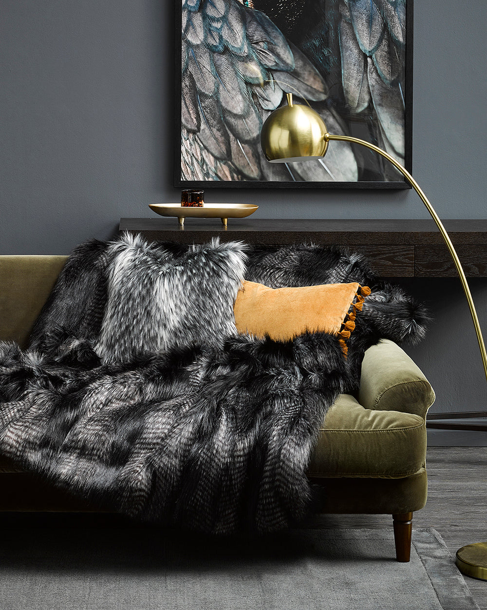 Alaskan Wolf imitation fur throw from heirloom in a lifestyle shote, grey scene with bird picture on the wall and Alaskan Wolf cushion on a green velvet sofa with a grey stone looking lamp and silver coffee table