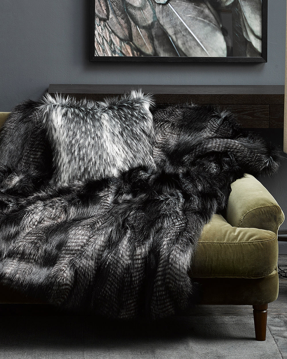 Alaskan Wolf imitation fur throw from heirloom in a lifestyle shote, grey scene with bird picture on the wall and Alaskan Wolf cushion on a green velvet sofa with a grey stone looking lamp and silver coffee table