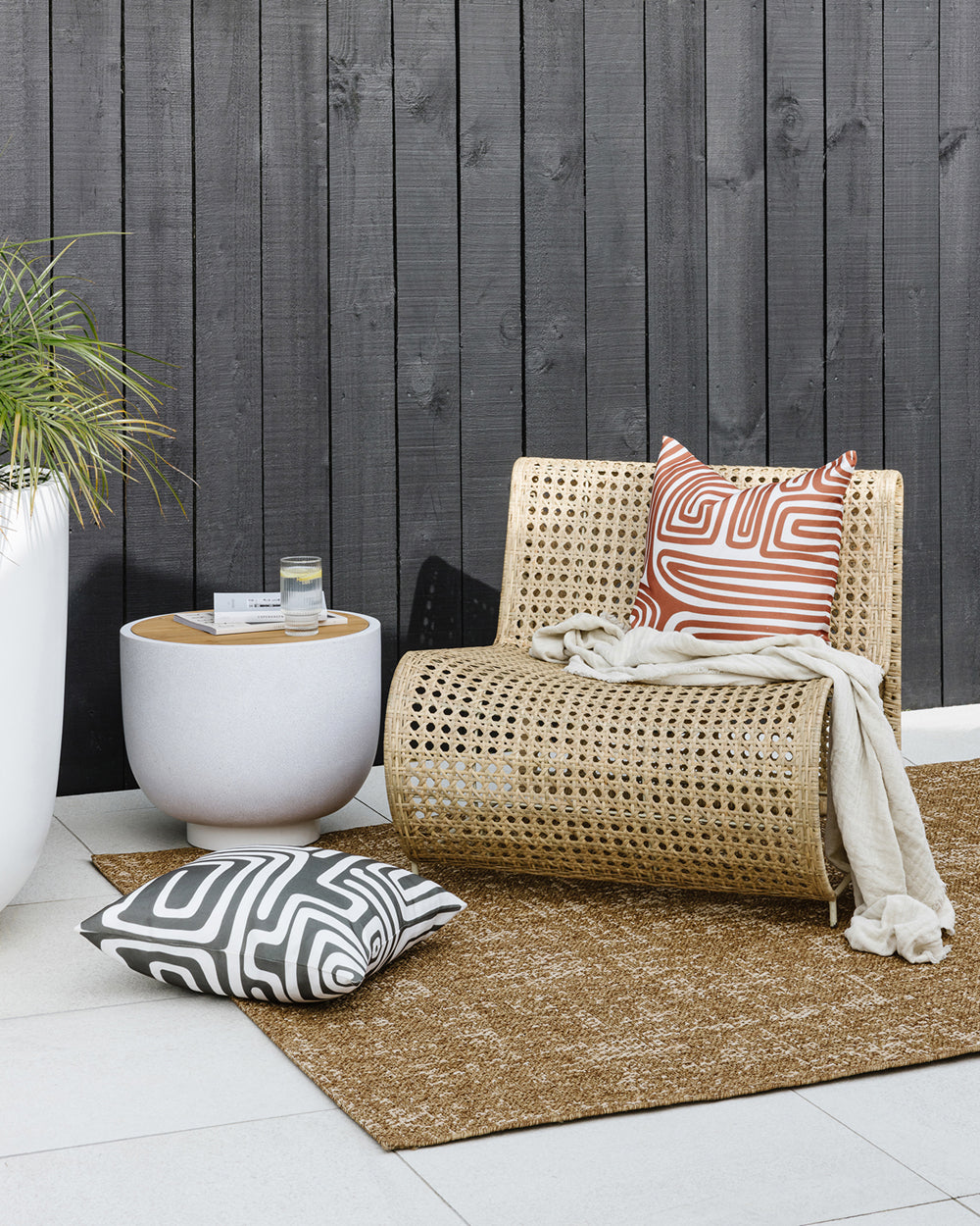 Ano outdoor cushion, rust abstract lines on the Ano on a rattan seat in an outdoor setting. Black cladding wooden wall with pico cushion on a taupe outdoor rug, concrete side table with drinks and a concrete large pot with greenery