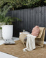 Ano outdoor cushion, rust abstract lines on the Ano on a rattan seat in an outdoor setting. Black cladding wooden wall with pico cushion on a taupe outdoor rug, concrete side table with drinks and a concrete large pot with greenery, green hedge behind the black cladded fence